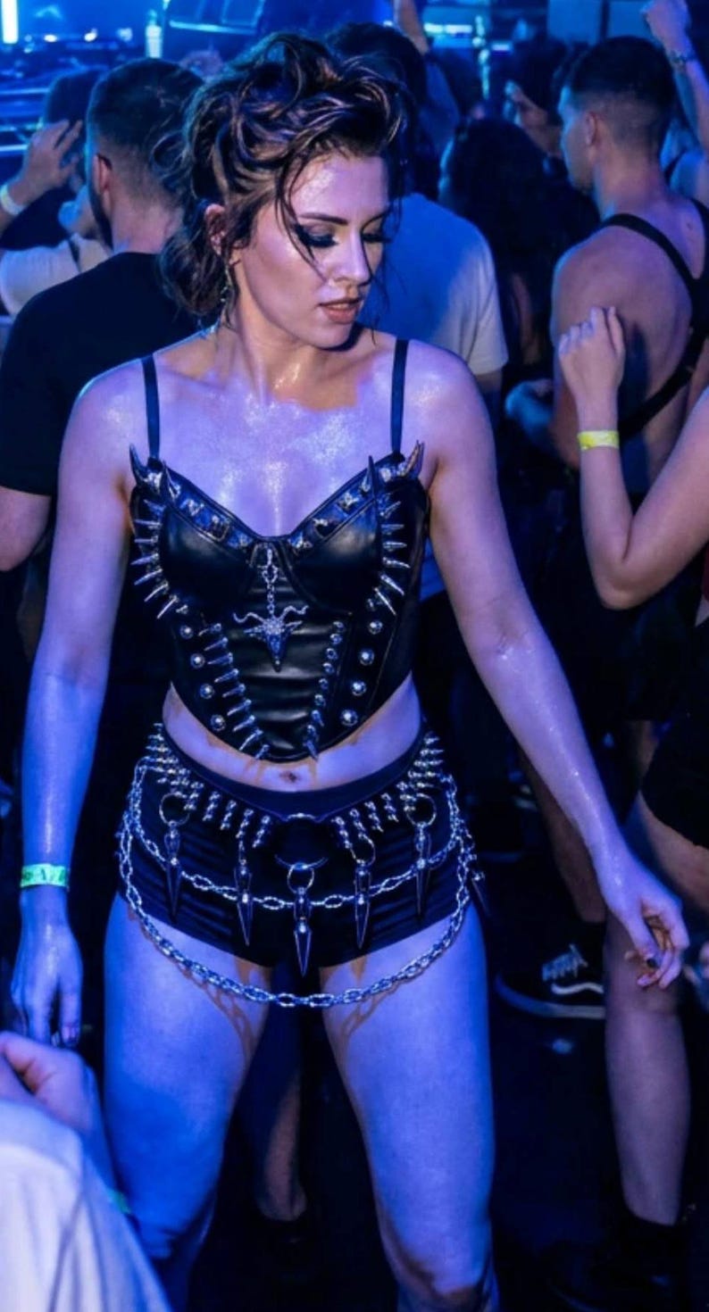 May include: A person in a crowded venue wearing a black leather corset with silver spikes and a matching belt with chain details. The outfit is accessorized with a chain belt and black shorts. The scene is illuminated with blue lighting.