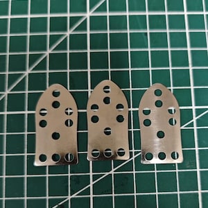 Puede incluir: Tres plantillas de diapasón de guitarra de metal plateado con múltiples agujeros y una parte superior redondeada. Las plantillas están dispuestas sobre una esterilla de corte verde con un patrón de cuadrícula.
