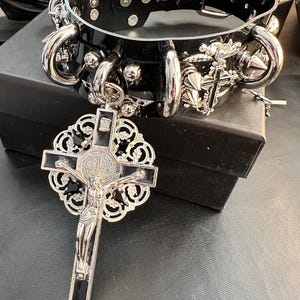 Puede incluir: Un collar de cuero negro con detalles en metal plateado, incluyendo pinchos, anillos y un colgante de cruz. El colgante presenta un dise&ntilde;o detallado y un fondo de esmalte negro.