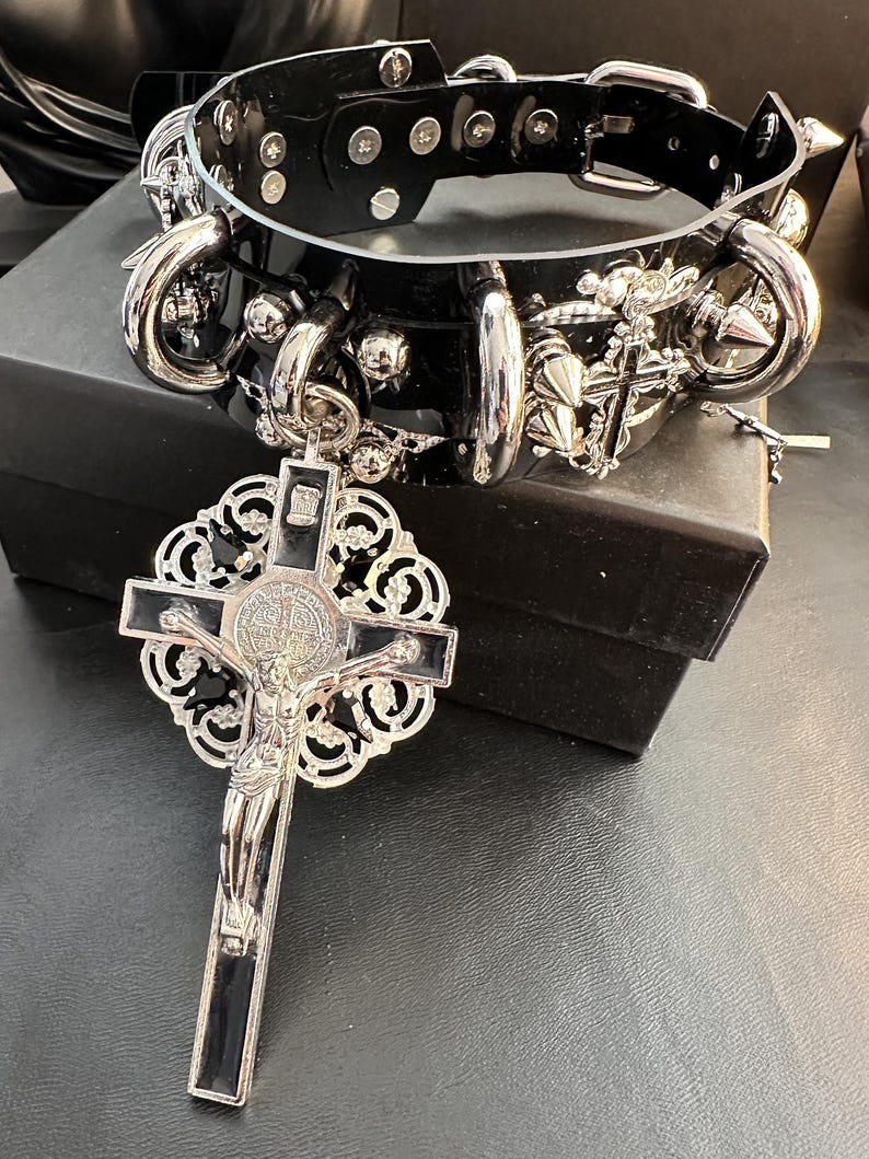 Puede incluir: Un collar de cuero negro con detalles en metal plateado, incluyendo pinchos, anillos y un colgante de cruz. El colgante presenta un dise&ntilde;o detallado y un fondo de esmalte negro.