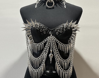 Peut inclure: Un corset en similicuir noir orné de pointes métalliques argentées sur le buste et de multiples chaînes argentées drapées avec des breloques en forme de pointe et un pendentif crâne central, assorti d'un tour de cou à pointes avec des croix.