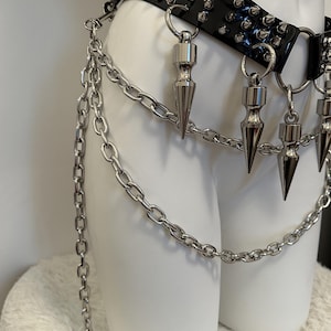 May include: A black, studded belt with silver-colored spikes and chain accents. The belt features a glossy finish and is adorned with multiple dangling, cone-shaped pendants. The chain hangs down, adding a layered effect to the design.