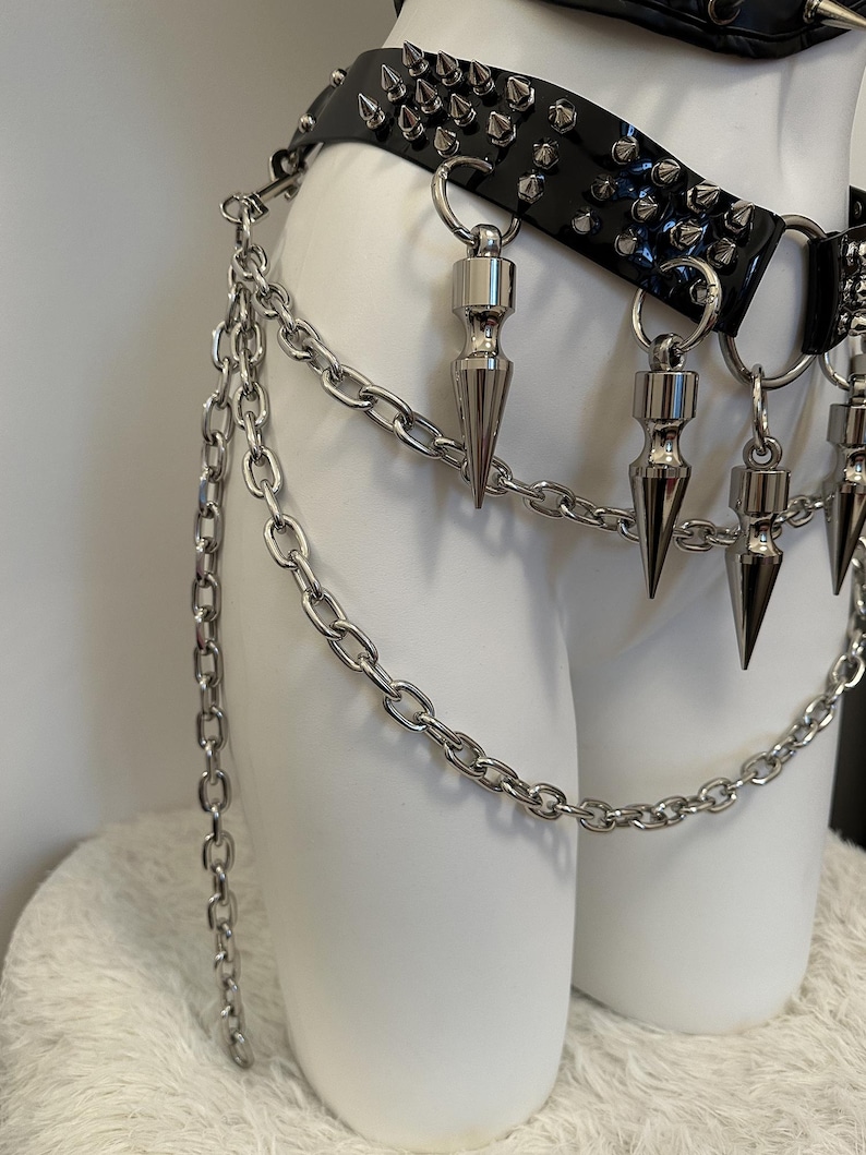 May include: A black, studded belt with silver-colored spikes and chain accents. The belt features a glossy finish and is adorned with multiple dangling, cone-shaped pendants. The chain hangs down, adding a layered effect to the design.