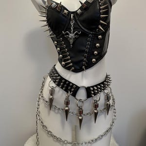 May include: Black faux leather corset top and belt set with silver-tone metal spikes and chain accents. The corset features a bull pendant and adjustable straps. The belt has dangling cone-shaped spikes and a chain that hangs down.