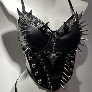 May include: Black faux leather corset with silver-toned spikes and studs. The corset features a decorative pendant with a horned skull design and adjustable straps. The garment is displayed on a white mannequin.