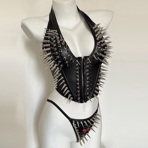 May include: A black halter-style corset top and matching thong set featuring numerous silver-toned metal spikes arranged in vertical rows across the front. The garment includes a central front closure and a small red skull detail on the thong.