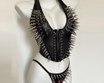 May include: A black halter-style corset top and matching thong set featuring numerous silver-toned metal spikes arranged in vertical rows across the front. The garment includes a central front closure and a small red skull detail on the thong.