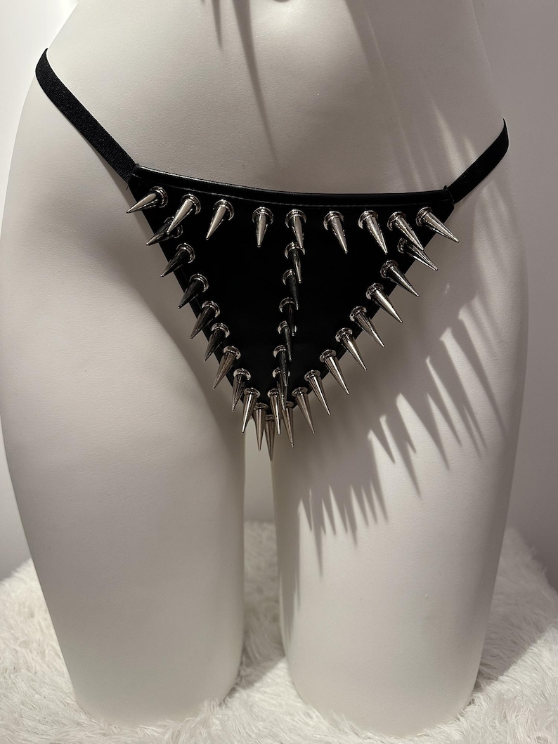 May include: Black leather thong with silver-colored metal spikes. The thong features a triangular design with multiple rows of spikes. The garment is displayed on a white mannequin.