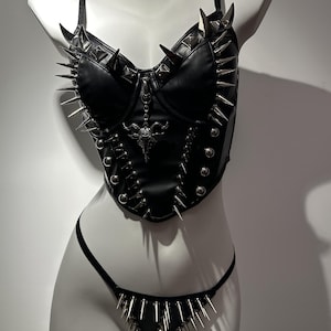 May include: Black leather corset and thong set with silver spikes and studs. The corset features a bull skull pendant and adjustable straps. The thong has a triangular front with matching spikes. The set is displayed on a white mannequin.