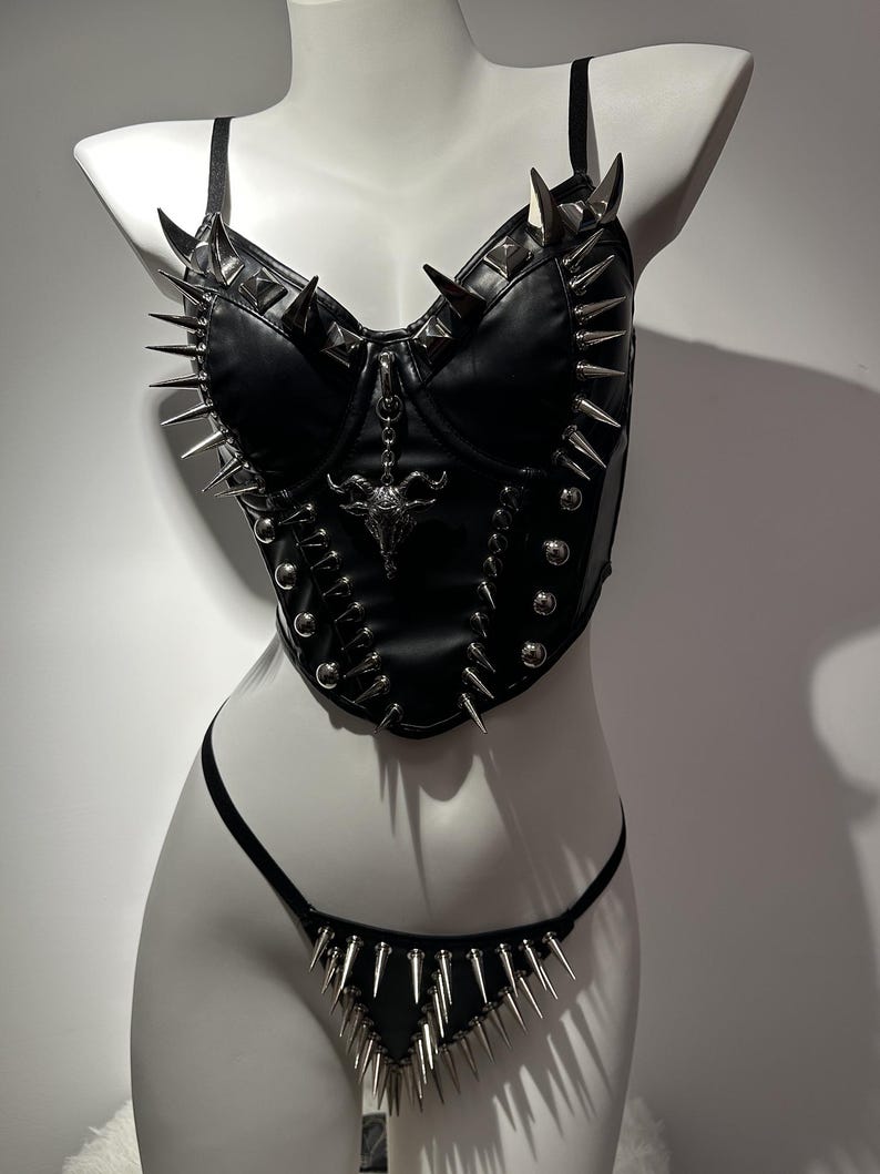 May include: Black leather corset and thong set with silver spikes and studs. The corset features a bull skull pendant and adjustable straps. The thong has a triangular front with matching spikes. The set is displayed on a white mannequin.