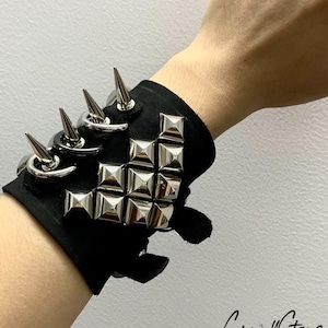 Puede incluir: Brazalete de muñeca de cuero negro con tachuelas piramidales y púas plateadas. El brazalete es ajustable y tiene un cierre de velcro.