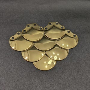 May include: A collection of shiny, gold-colored metal scales arranged in a layered pattern. Each scale is a half-circle shape with small holes at the top. The scales are on a dark gray surface.
