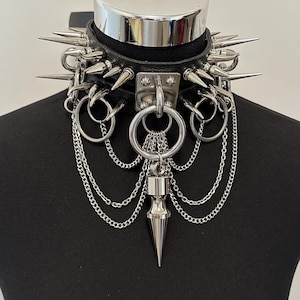 May include: A black leather choker collar featuring silver-toned metal spikes, multiple circular rings, and layered silver chains. A central square metal plate holds a large ring with a dangling silver-toned metal spike pendant at the bottom.