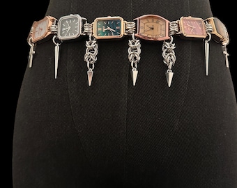 May include: A unique belt featuring multiple rectangular watch faces in various colors and metal finishes. The watches are connected by silver-toned wire and adorned with dangling silver-toned chain and spike charms. The belt is displayed on a black mannequin.
