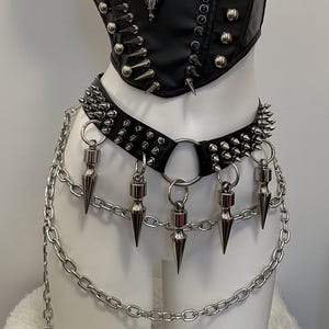 May include: Black leather corset and belt set with silver spikes and chain details. The corset features a pendant with a horned skull. The belt has dangling cone-shaped charms and a chain that hangs down.