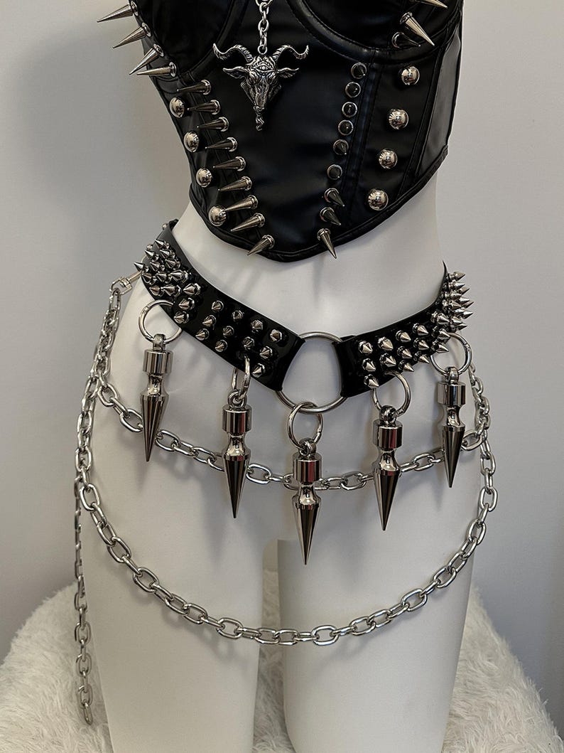 May include: Black leather corset and belt set with silver spikes and chain details. The corset features a pendant with a horned skull. The belt has dangling cone-shaped charms and a chain that hangs down.