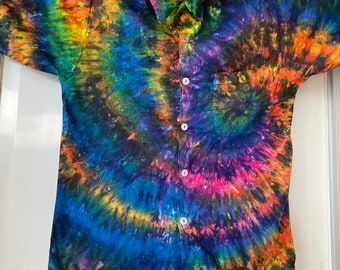 May include: A short-sleeved button-down shirt with a vibrant tie-dye pattern. The shirt features a swirling design of blues, greens, yellows, oranges, and purples. It has a single chest pocket and white buttons. The shirt is displayed on a wooden hanger.