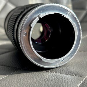 May include: Close-up of a black camera lens with a silver-colored mounting ring. The lens has a ridged focus ring and a clear aperture. The lens is designed for use with a camera.