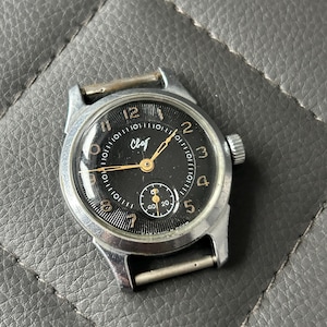 May include: A vintage wristwatch with a black face, silver hands, and a silver case. The watch face has Arabic numerals and a small seconds dial. The watch has a textured crown and metal band attachments.
