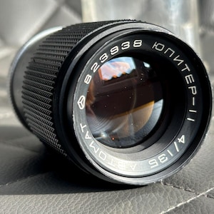 Può includere: Un obiettivo per fotocamera nero con il testo "Jupiter-11" e "823938" stampato sopra. L'obiettivo ha un'impugnatura strutturata e un elemento in vetro riflettente. L'obiettivo è di 13,5 cm.