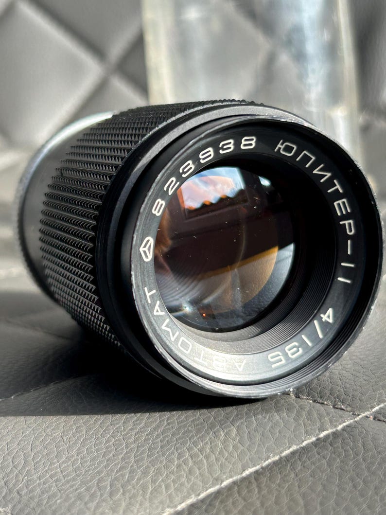 May include: A black camera lens with the text "Jupiter-11" and "823938" printed on the lens. The lens has a textured grip and a reflective glass element. The lens is 135mm.