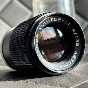May include: Black camera lens with the text "ЮПИТЕР-11" and "4/135 ABTOMAT" in white lettering. The lens has a textured grip and a clear protective cover in the background. The lens is designed for photography.