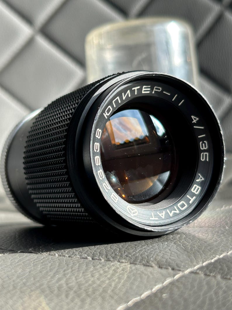 May include: Black camera lens with the text "ЮПИТЕР-11" and "4/135 ABTOMAT" in white lettering. The lens has a textured grip and a clear protective cover in the background. The lens is designed for photography.