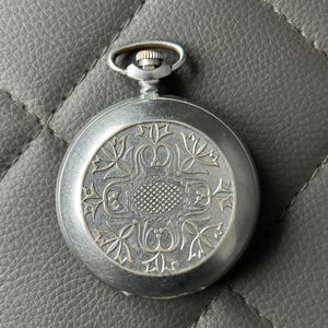 Puede incluir: Un reloj de bolsillo plateado con un diseño floral ornamentado en la tapa. El reloj tiene una esfera circular con un patrón texturizado en el centro y un lazo en la parte superior para una cadena. El reloj está sobre un fondo gris.