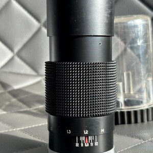 May include: Black camera lens with a textured grip and a silver and white adjustment ring. The lens has a series of numbers and markings, including f-stop indicators. A clear plastic lens cap is visible in the background.
