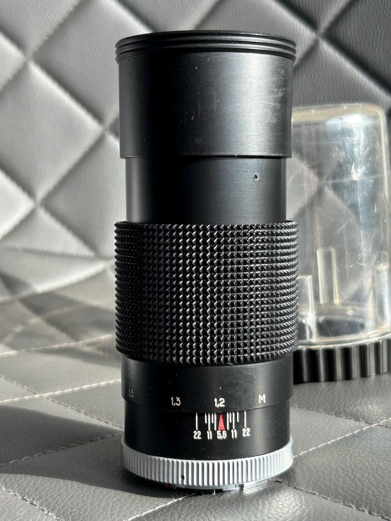 May include: Black camera lens with a textured grip and a silver and white adjustment ring. The lens has a series of numbers and markings, including f-stop indicators. A clear plastic lens cap is visible in the background.