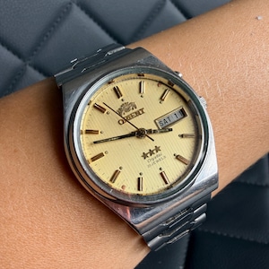 May include: A silver-tone analog wristwatch with a textured gold dial, featuring the Orient logo, three stars, and the text Crystal 21 Jewels. The watch includes a day-date window displaying SAT 11, gold-tone hour markers, and a metal link bracelet.