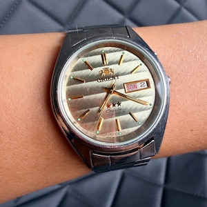 May include: An Orient wristwatch with a silver-tone metal link band and a round champagne-colored dial featuring horizontal textured stripes. The watch face displays gold-tone hands, hour markers, a day-date window showing SON 2, and the text Orient Crystal 21 Jewels.