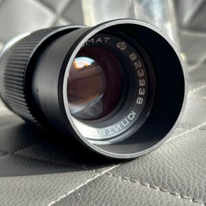 May include: A black camera lens with the text "MAT" and "823938" printed on the lens. The lens has a textured grip and a reflective glass element. The lens is cylindrical in shape.