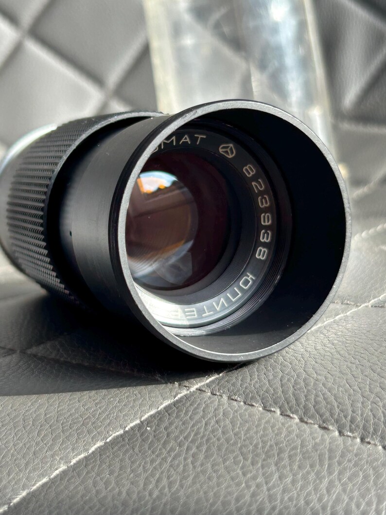 May include: A black camera lens with the text "MAT" and "823938" printed on the lens. The lens has a textured grip and a reflective glass element. The lens is cylindrical in shape.