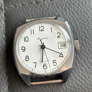 Könnte beinhalten: Eine Vintage Raketa Armbanduhr mit einem quadratischen silberfarbenen Gehäuse und einem weißen Zifferblatt. Die Uhr hat schwarze Zeiger, arabische Ziffern und ein Datumsfenster. Die Wörter "Raketa" und "Made in USSR" sind sichtbar. Das Gehäuse ist ca. 3,5 cm breit.