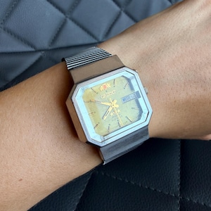May include: A silver-toned Orient wristwatch with an octagonal case and a textured gold-colored dial. The watch features gold-colored hands, hour markers, and a day-date window at the three o'clock position. It includes a flexible metal link band.