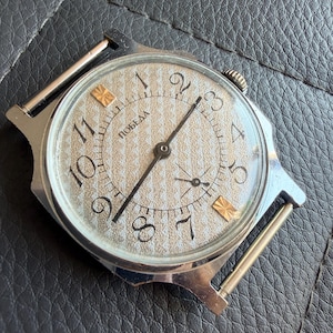 May include: A vintage Pobeda wristwatch featuring a silver-toned case and a textured, light-coloured dial with black Arabic numerals. The dial displays the Cyrillic text 'ПОБЕДА', thin black hands, a small sub-dial for seconds, and two gold-toned markers.