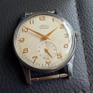 May include: A vintage Prim wristwatch with a textured cream-colored dial, gold-toned Arabic numerals, and matching gold-toned hands. The watch features a small sub-dial for seconds at the six o'clock position and a silver-toned metal case.