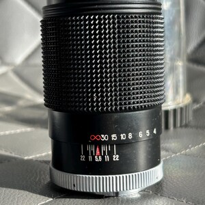 May include: A black camera lens with a textured grip and silver accents. The lens has a series of numbers and markings, including 22, 11, 5.6, 11, and 22. A clear plastic lens cap is visible in the background.