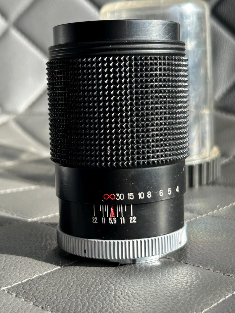 May include: A black camera lens with a textured grip and silver accents. The lens has a series of numbers and markings, including 22, 11, 5.6, 11, and 22. A clear plastic lens cap is visible in the background.