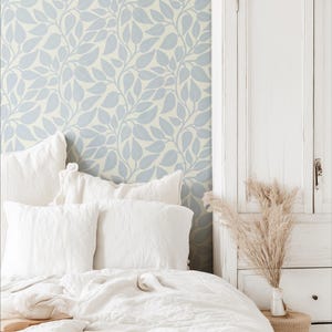 May include: A bedroom scene featuring a bed with white linens and pillows. The headboard wall has a light blue and white leaf pattern wallpaper. A white cabinet and a small woven stool with a vase of dried plants complete the scene.