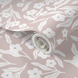 May include: A roll of wallpaper featuring a repeating pattern of white flowers and leaves on a muted pink background. The floral design is stylized and creates a soft, inviting aesthetic. The wallpaper is partially unrolled, revealing the pattern.