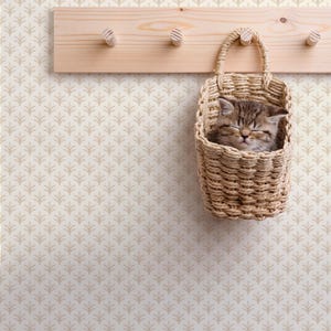 May include: A tabby kitten rests peacefully in a woven basket hanging from a wooden peg rack. The basket is made of natural fibers and has a handle. The background is a cream-colored wall with a repeating leaf pattern.
