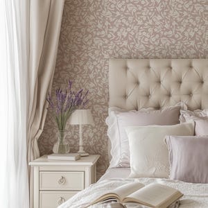 May include: A bedroom scene featuring a tufted headboard, pillows, and a bedspread. A nightstand holds a lamp, a vase of lavender, and a book. The wallpaper has a floral pattern, and curtains frame the window.