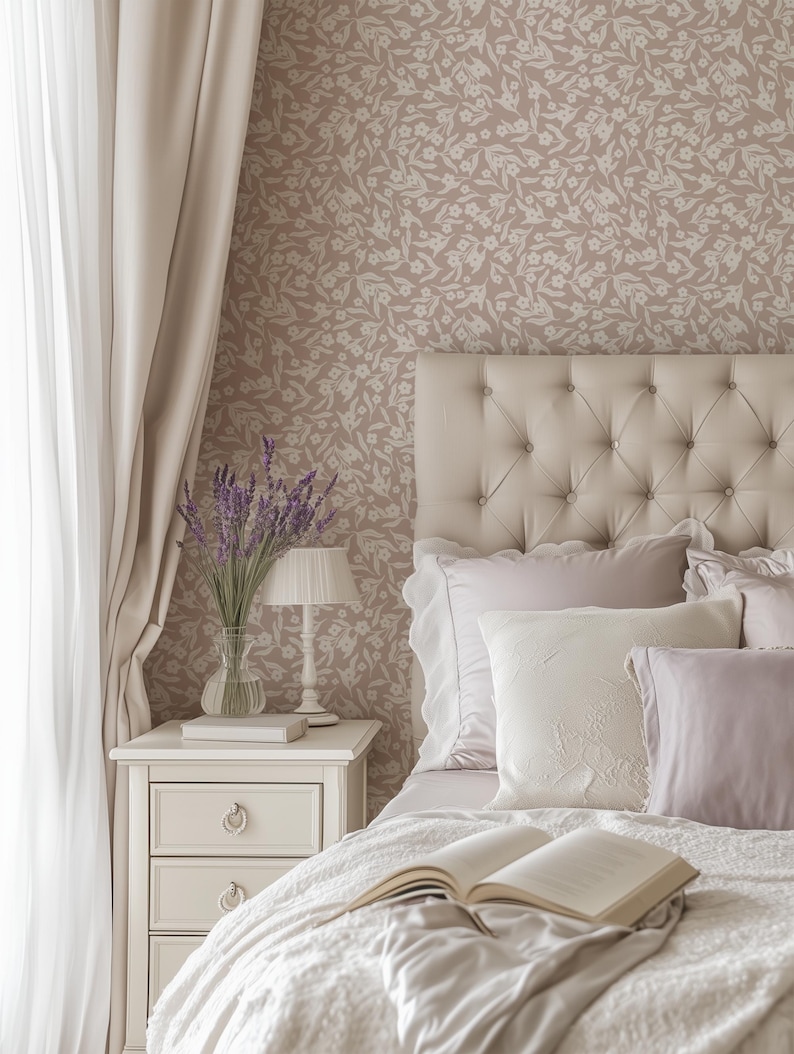 May include: A bedroom scene featuring a tufted headboard, pillows, and a bedspread. A nightstand holds a lamp, a vase of lavender, and a book. The wallpaper has a floral pattern, and curtains frame the window.