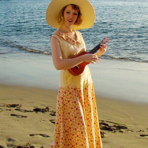 May include: A woman wearing a yellow floral dress and a wide-brimmed straw hat plays a ukulele on a sandy beach.