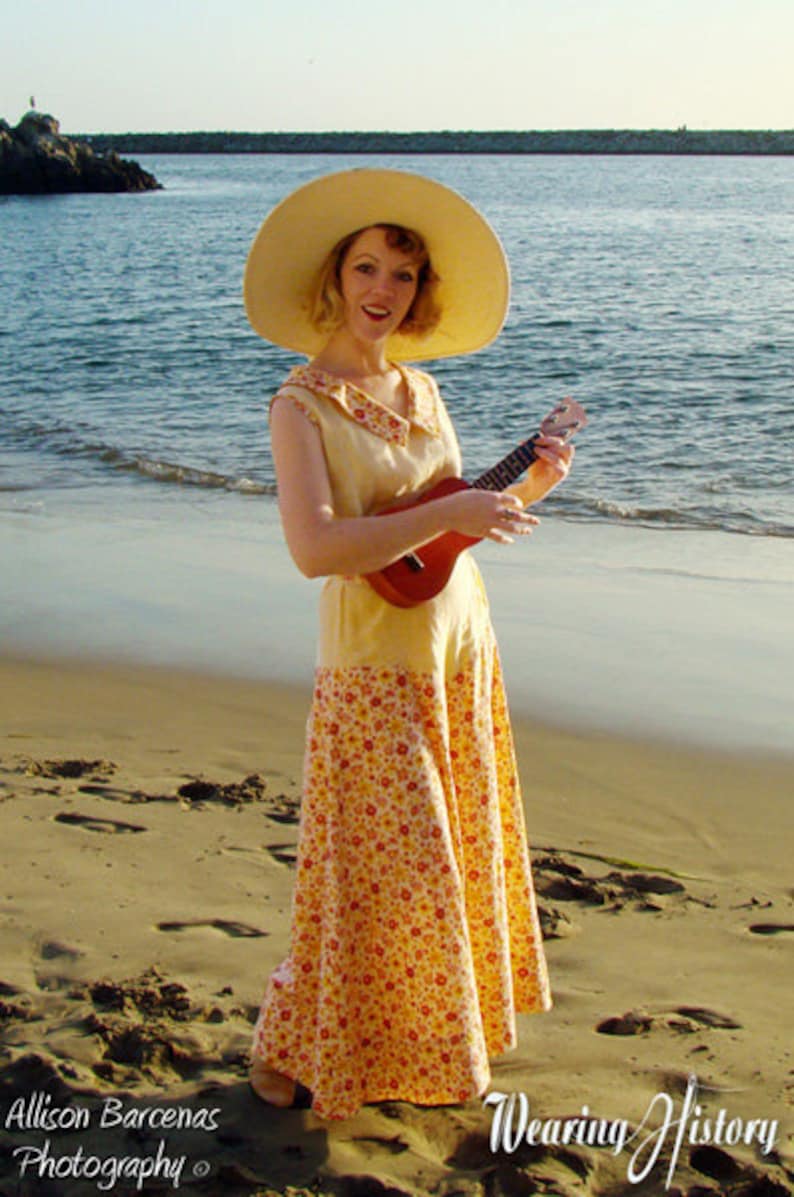 May include: A woman wearing a yellow floral dress and a wide-brimmed straw hat plays a ukulele on a sandy beach.