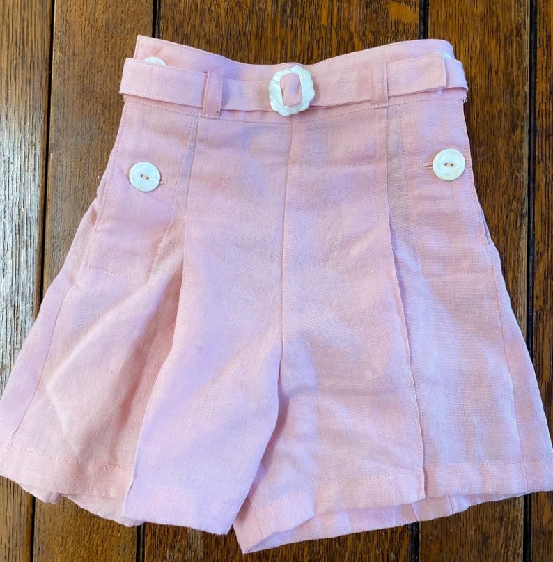 Może przedstawiać: R&oacute;żowe spodenki lniane z białym paskiem i guzikami. Spodenki mają fałdy z przodu i kieszenie po bokach.