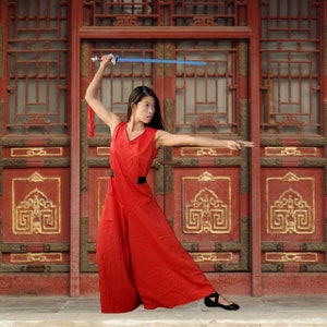 May include: A woman in a red jumpsuit with a black belt stands in front of a red wooden door with gold accents. She holds a blue sword in her right hand and is in a martial arts pose.