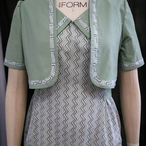 May include: A green cropped jacket with a white zig-zag trim over a white and gray chevron patterned dress. The dress has a white collar and a green trim around the neckline. The dress has a fitted bodice and a full skirt.